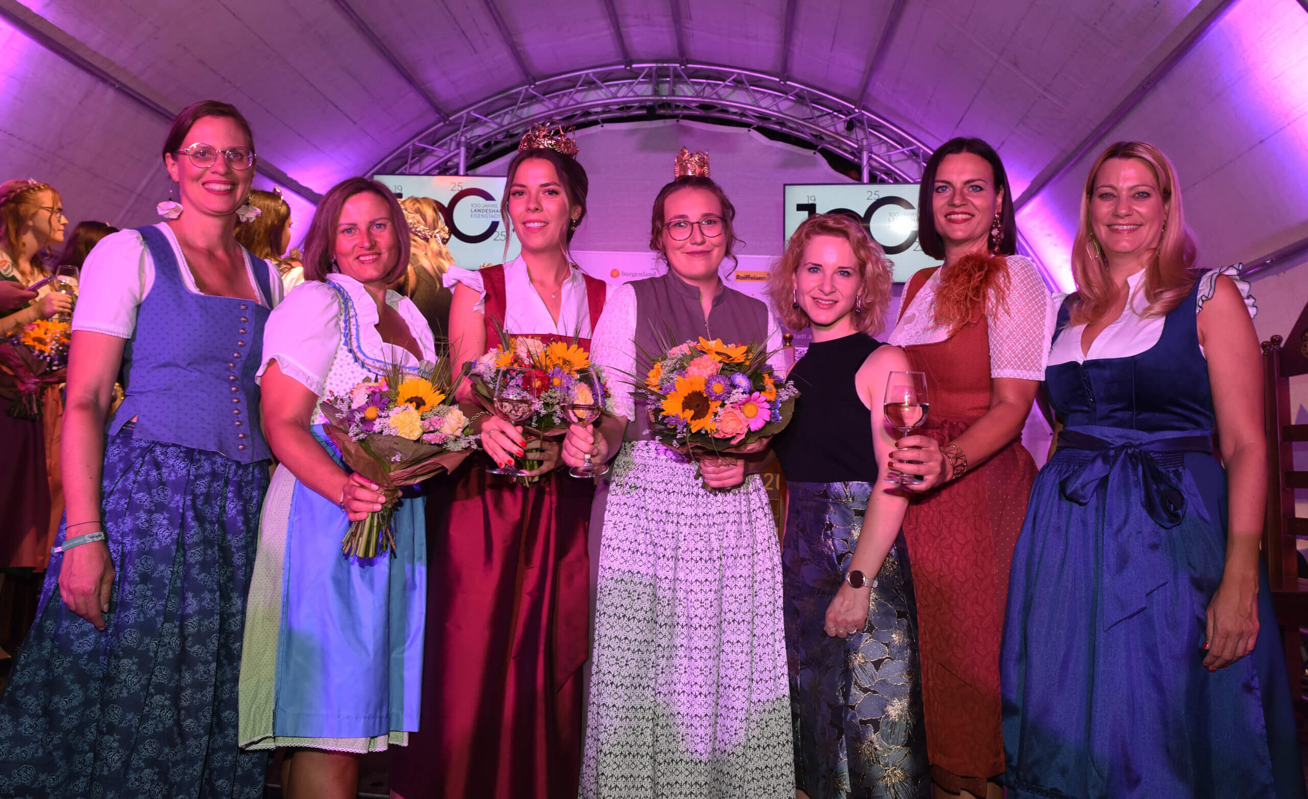 Gruppe von Frauen in traditionellen Dirndlkleidern bei einem festlichen Event mit Blumensträußen und Wein.