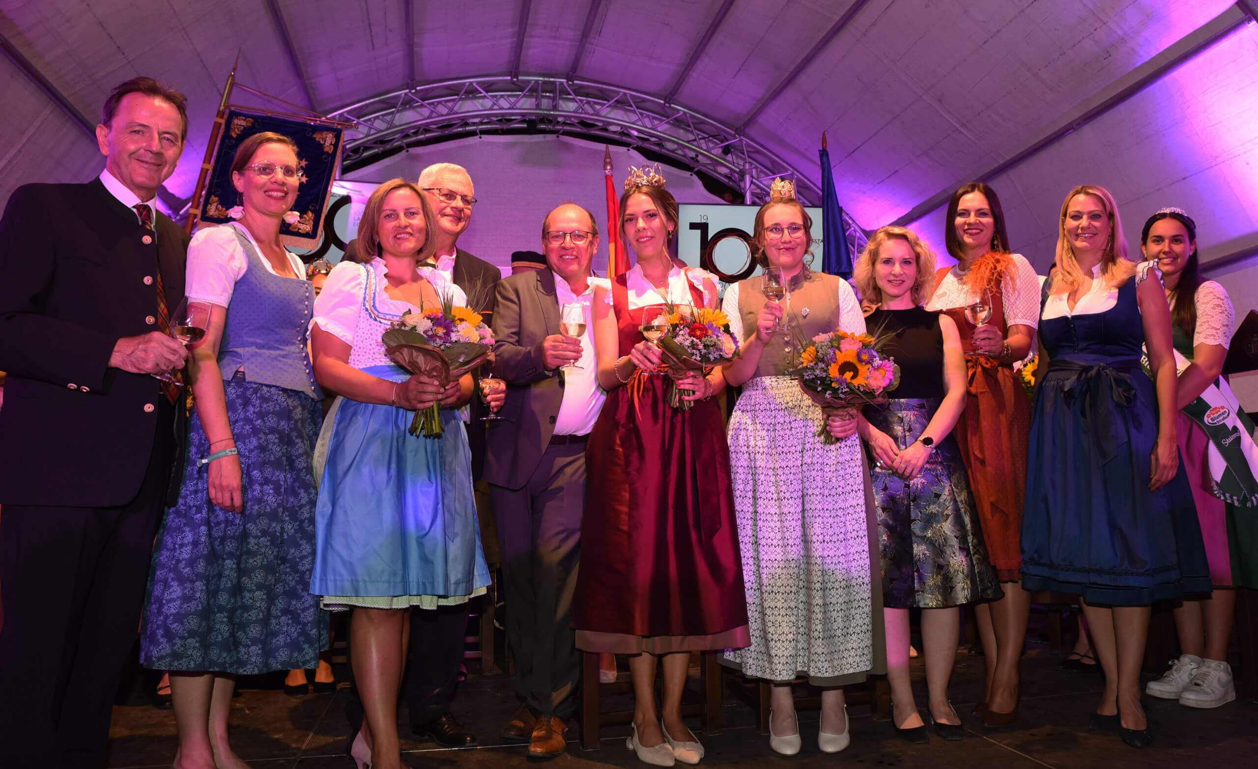 Personen in traditioneller Kleidung feiern mit Blumen und Sekt auf einem Fest. Fröhliche Gruppenaufnahme unter lila Licht.