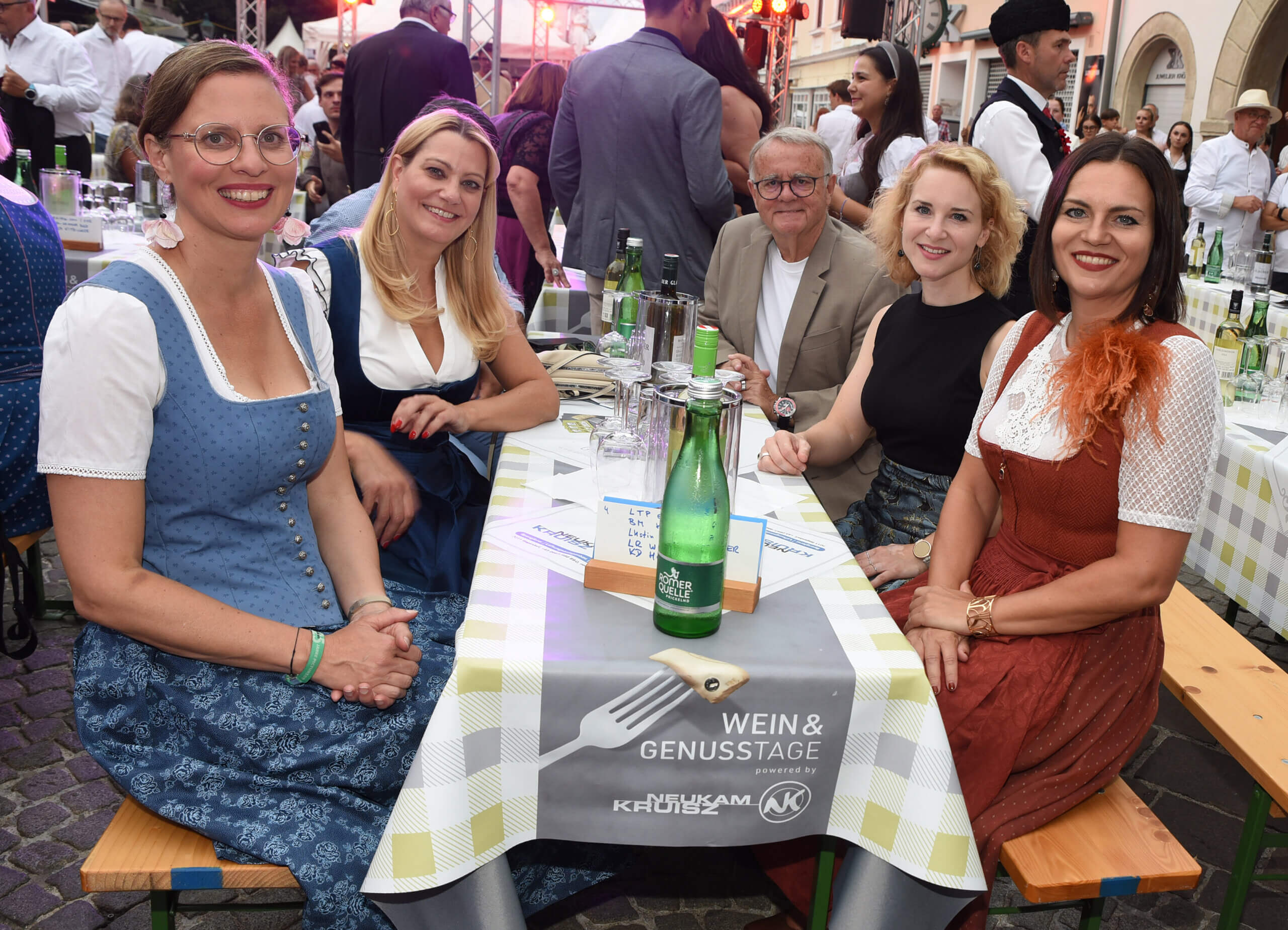 Gruppe von lächelnden Menschen in Tracht bei einem Outdoor-Event mit gedecktem Tisch und Getränken.