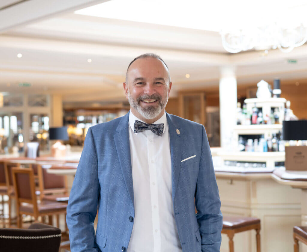 Gastgeber Peter Prisching©AVITA Resort Mann in blauem Anzug und Fliege lächelt in einem schicken Restaurantambiente.