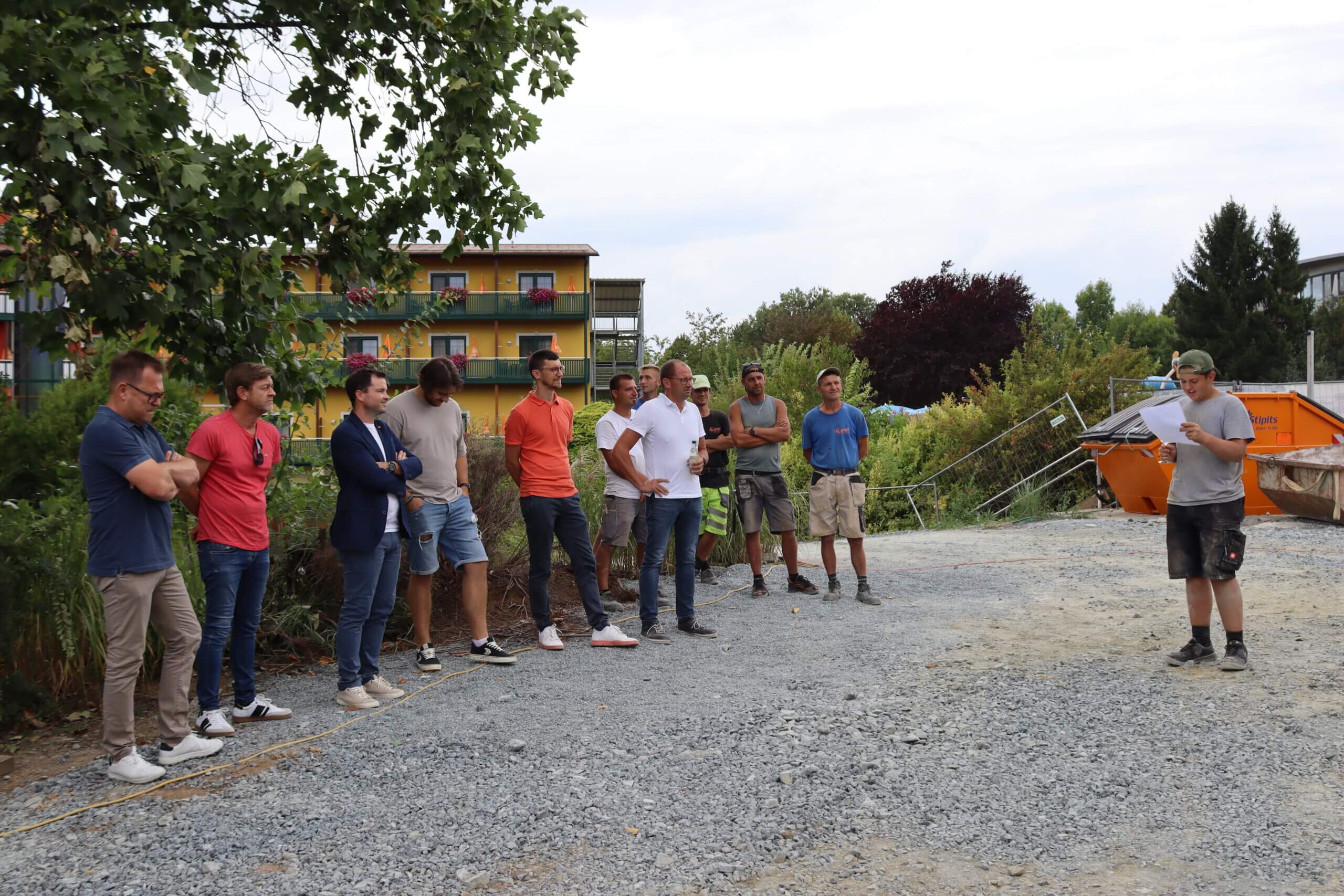 Gruppe von Menschen bei einer Baustellenbesprechung im Freien, mit Bäumen und einem bunten Gebäude im Hintergrund.