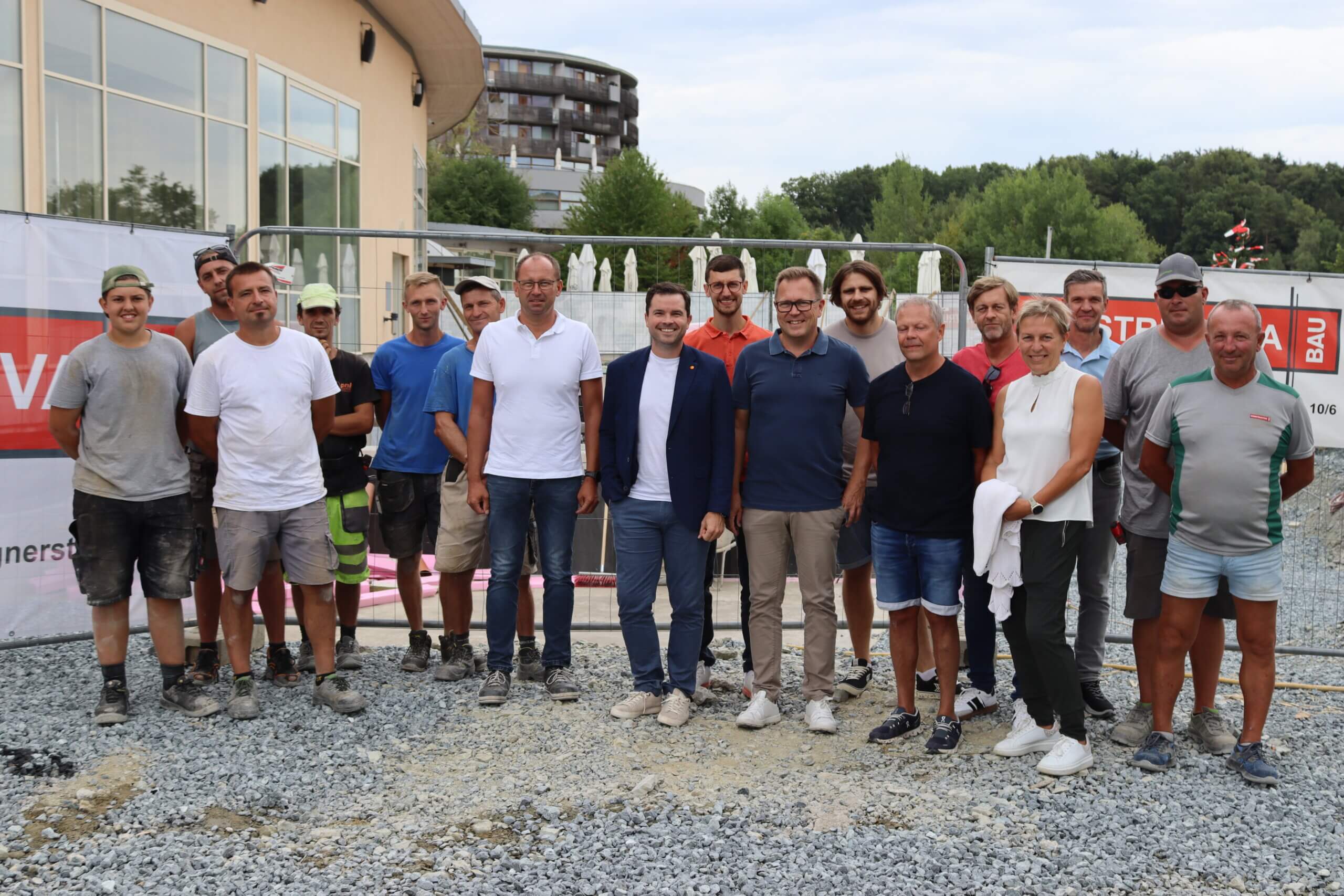 Gruppe von Bauarbeitern und Geschäftsleuten vor Baustelle, lächelnd in Arbeitskleidung und Freizeitkleidung.