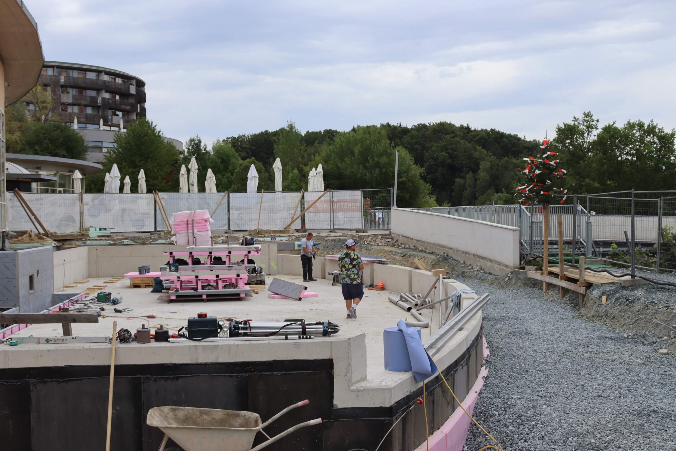 Bauarbeiten am Schwimmbad mit Richtbaum und Arbeitern, umgeben von modernen Gebäuden und grüner Natur.
