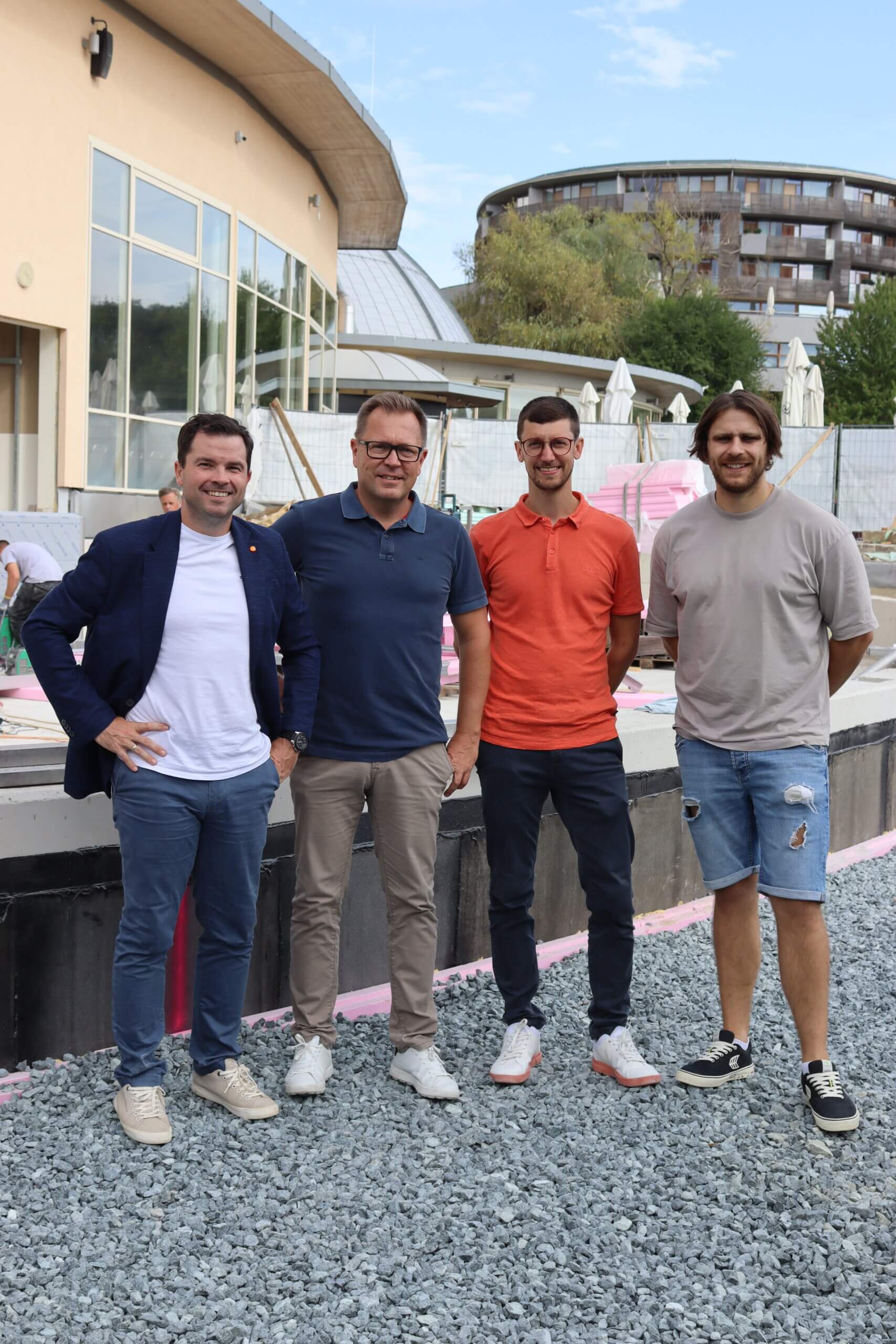 Vier Männer stehen auf einem Baustellengelände vor modernen Gebäuden unter blauem Himmel.