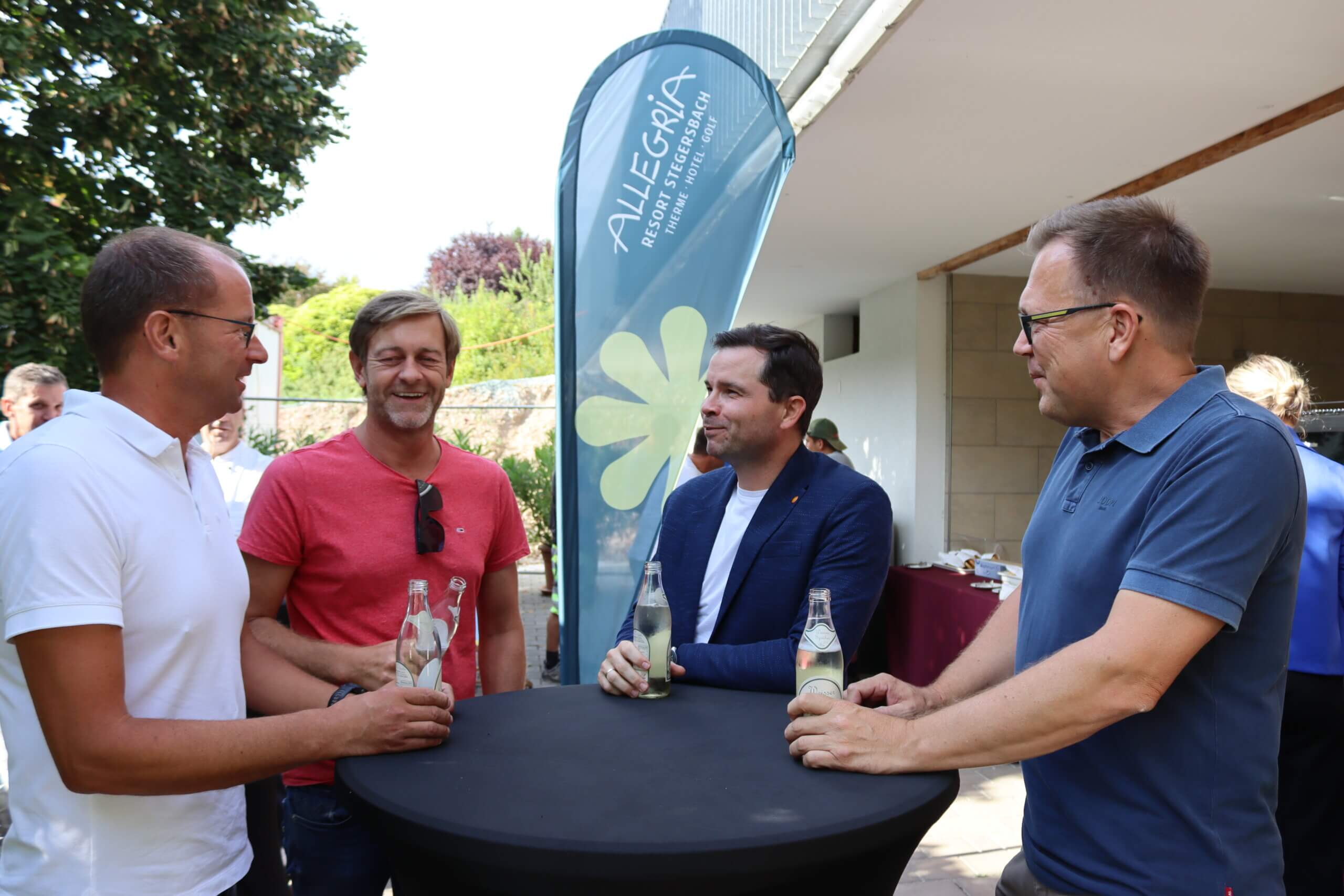 Vier Männer plaudern bei einem Event im Allegra Resort Stegersbach draußen am Tisch mit Getränken in der Hand.