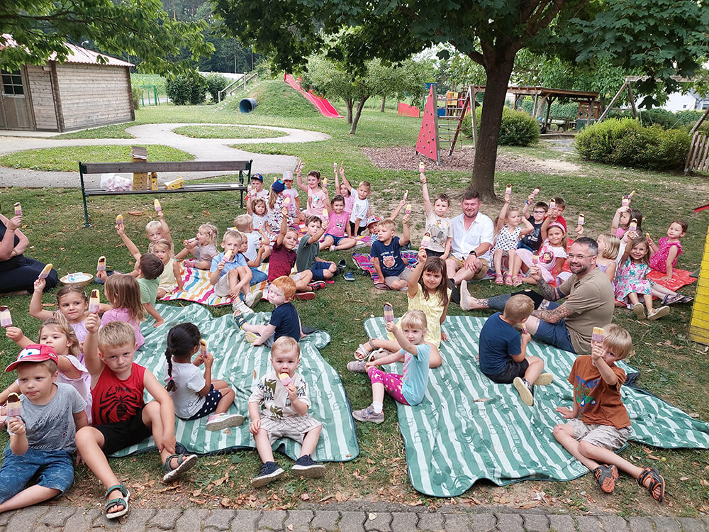 Gruppenfoto KiGa©Gemeinde Rohr bei Hartberg Kindergruppe genießt Eis im Freien, Sommerpicknick im Park mit vielen lachenden Kindern unter Bäumen.
