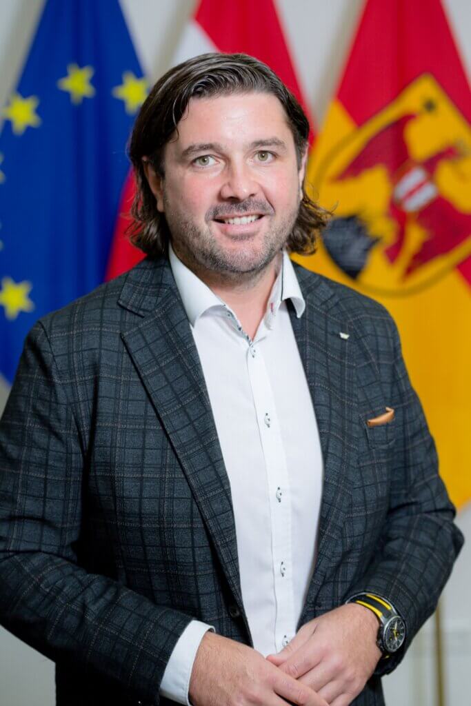 Ein Mann in einem karierten Anzug vor europäischen und nationalen Flaggen, lächelnd in die Kamera.