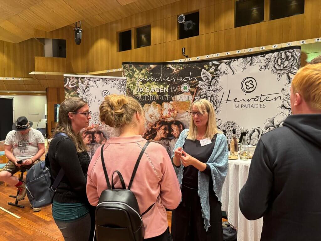 IMG-20241116-WA0014©Andreas Bruckner Gruppe diskutiert auf einer Hochzeitseventmesse im Sudburgenland, Stand Heiraten im Paradies, mit Informationsplakaten.