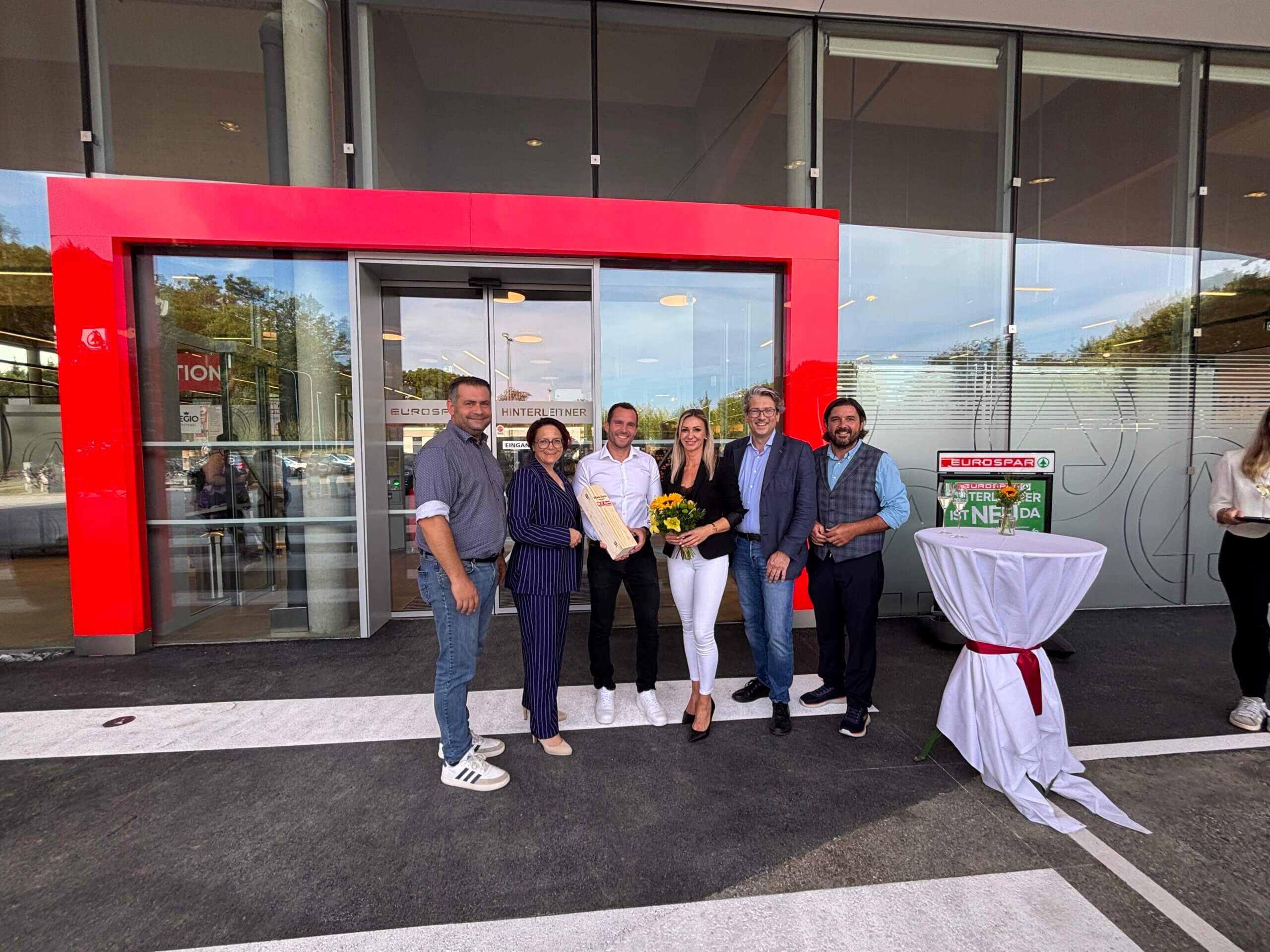 Gruppe von sechs Personen bei einer Geschäftseröffnung vor einem modernen Gebäude mit rotem Eingang.