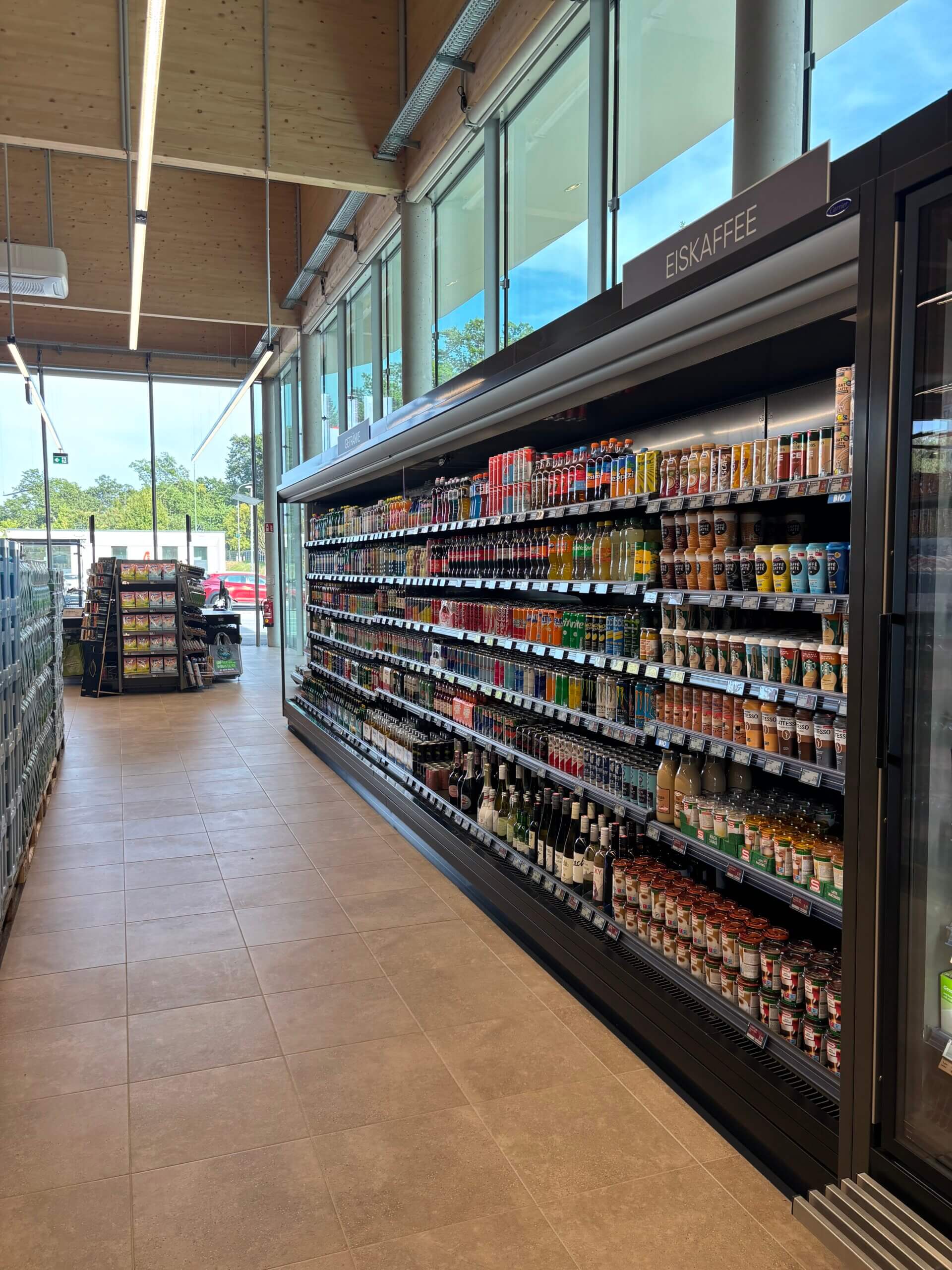 Supermarktgang mit Regalen voller Getränke und Eiskaffee, modernes Interieur, sonniger Tag draußen sichtbar durch Fenster.