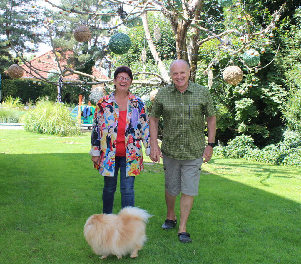 Paar mit Hund im Garten, bunt gekleidet, umgeben von dekorativen Bäumen und grünem Rasen bei sonnigem Wetter.
