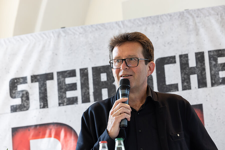 Ein Mann mit Mikrofon spricht vor einem Banner auf einer Veranstaltung.