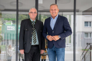 Zwei Männer in Anzügen lächeln vor einem Bürogebäude in die Kamera. Geschäftsbegegnung im Freien.