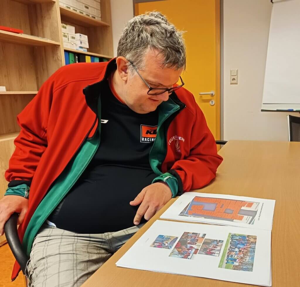 LH HB_Selbstvertretung_(c)Lebenshilfe Hartberg (3) Mann in roter Jacke blickt auf Dokumente an einem Tisch in einem Büro.