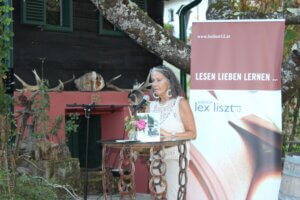 Frau hält Lesung im Freien neben Banner von Edition Lex Liszt mit Slogan Lesen Lieben Lernen.