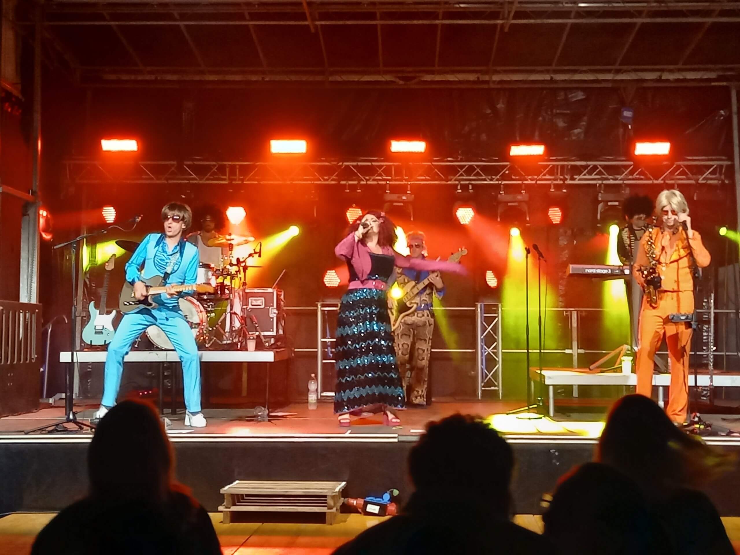Band in Retro-Outfits spielt live auf Bühne mit buntem Licht, Musiker mit Gitarre, Sängerin in Bewegung.