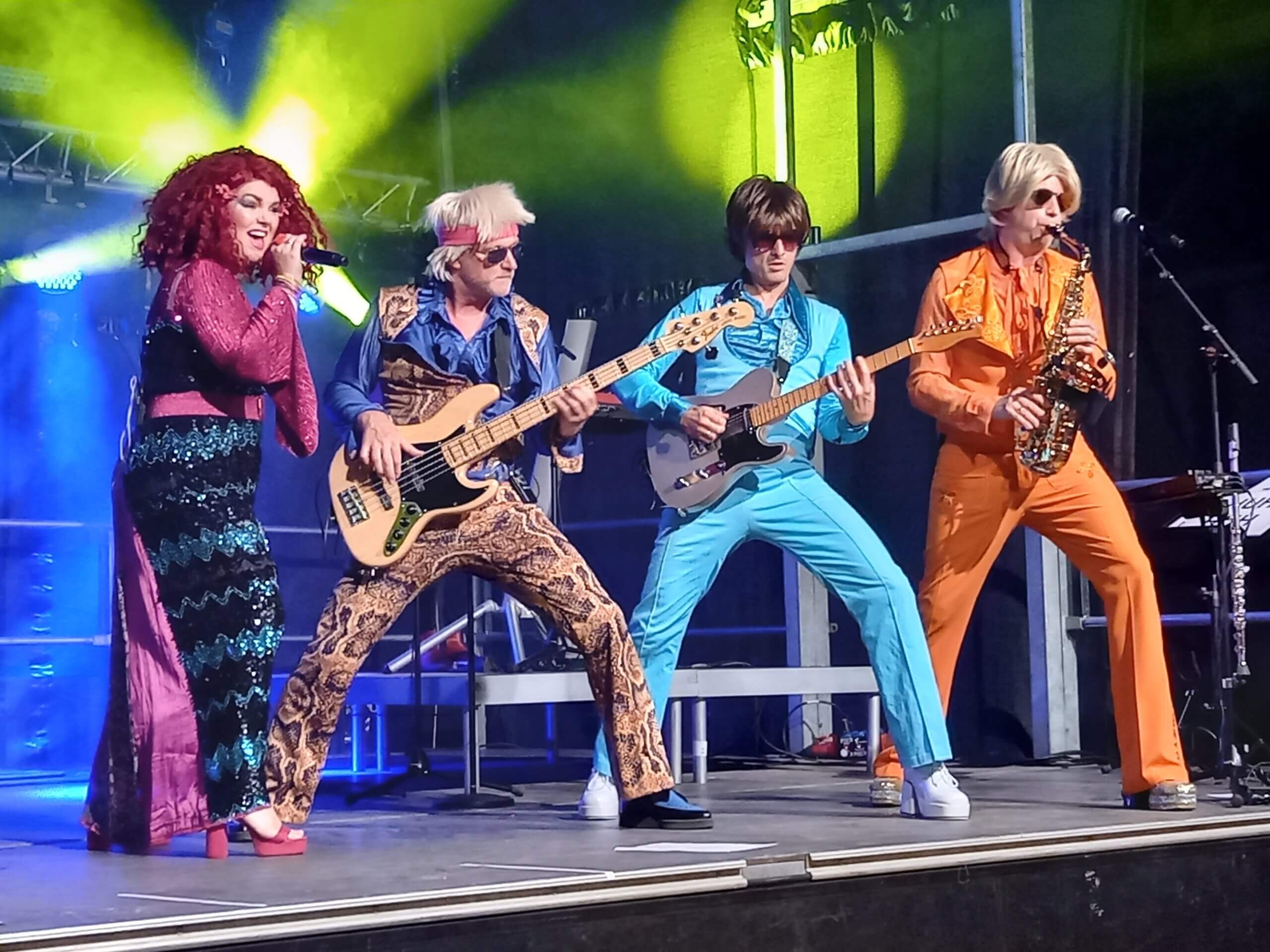 Band performt live auf Bühne mit bunten Kostümen und Instrumenten, energiegeladene Show, Beleuchtung im Hintergrund.