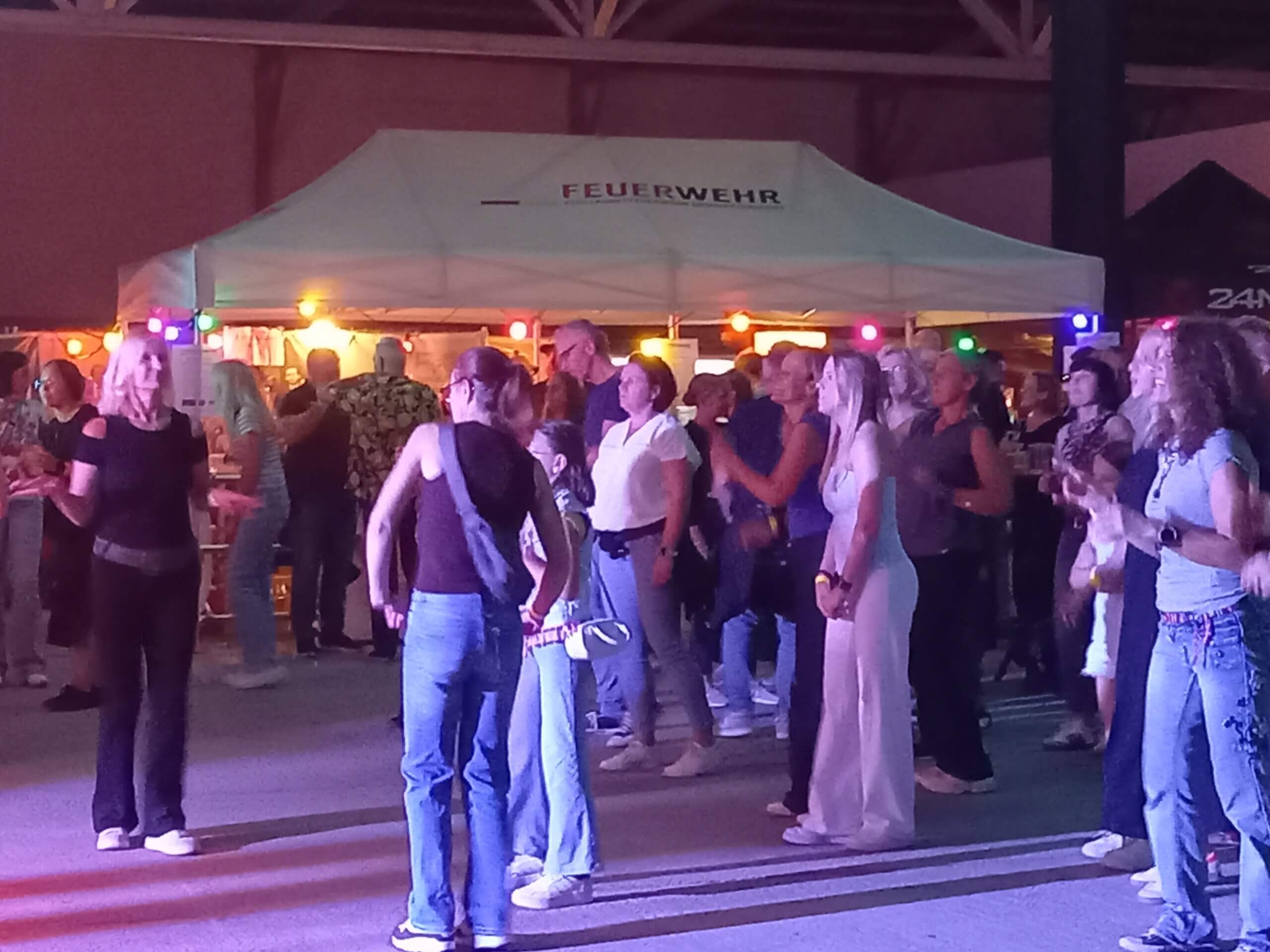 Menschen tanzen vor einem Feuerwehrzelt bei einer Veranstaltung in der Nacht, umgeben von bunten Lichtern.