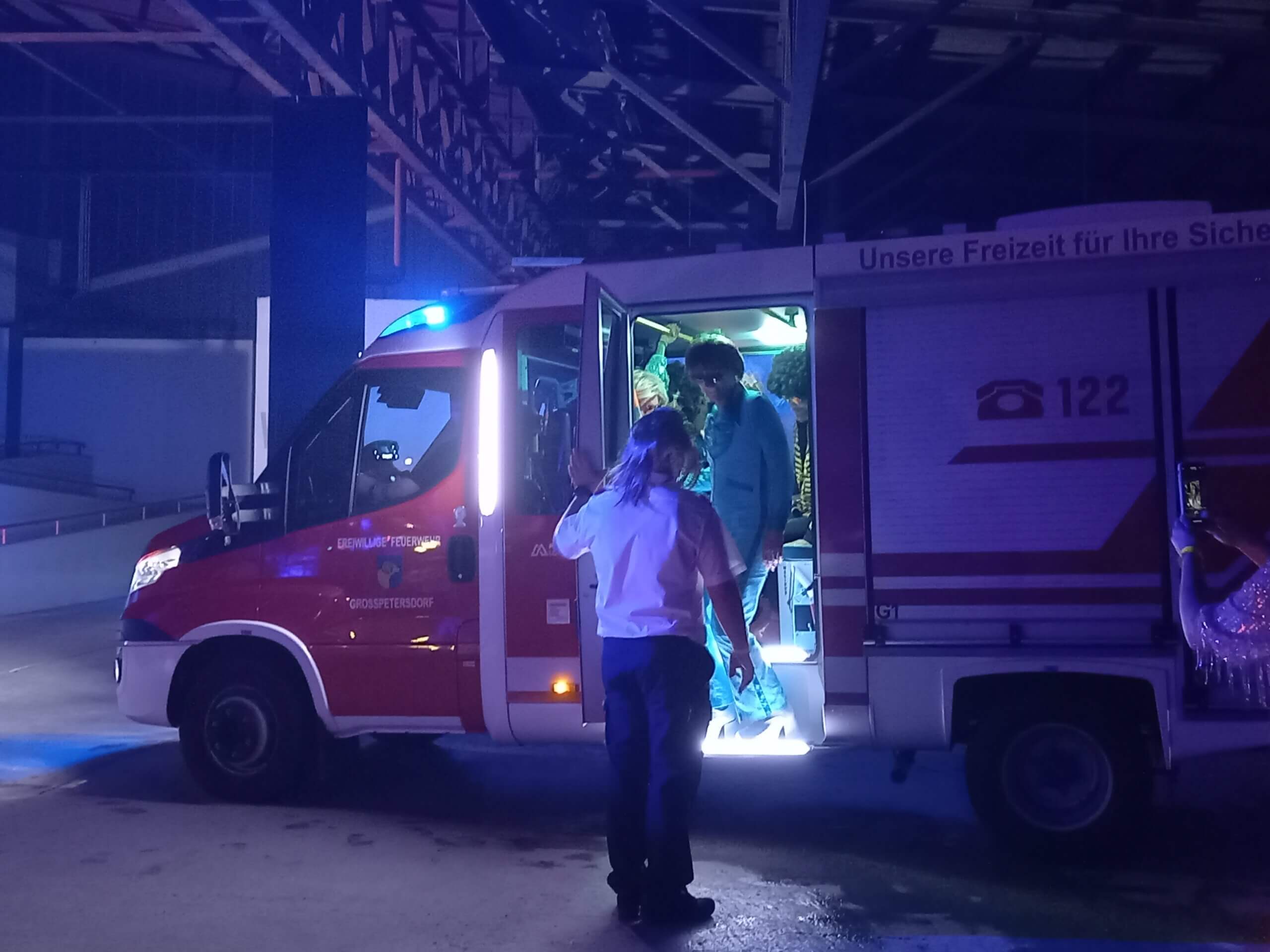 Feuerwehrwagen mit Personen bei Nacht in einer Halle, Blaulicht leuchtet auf, Freiwillige Feuerwehr im Einsatz.