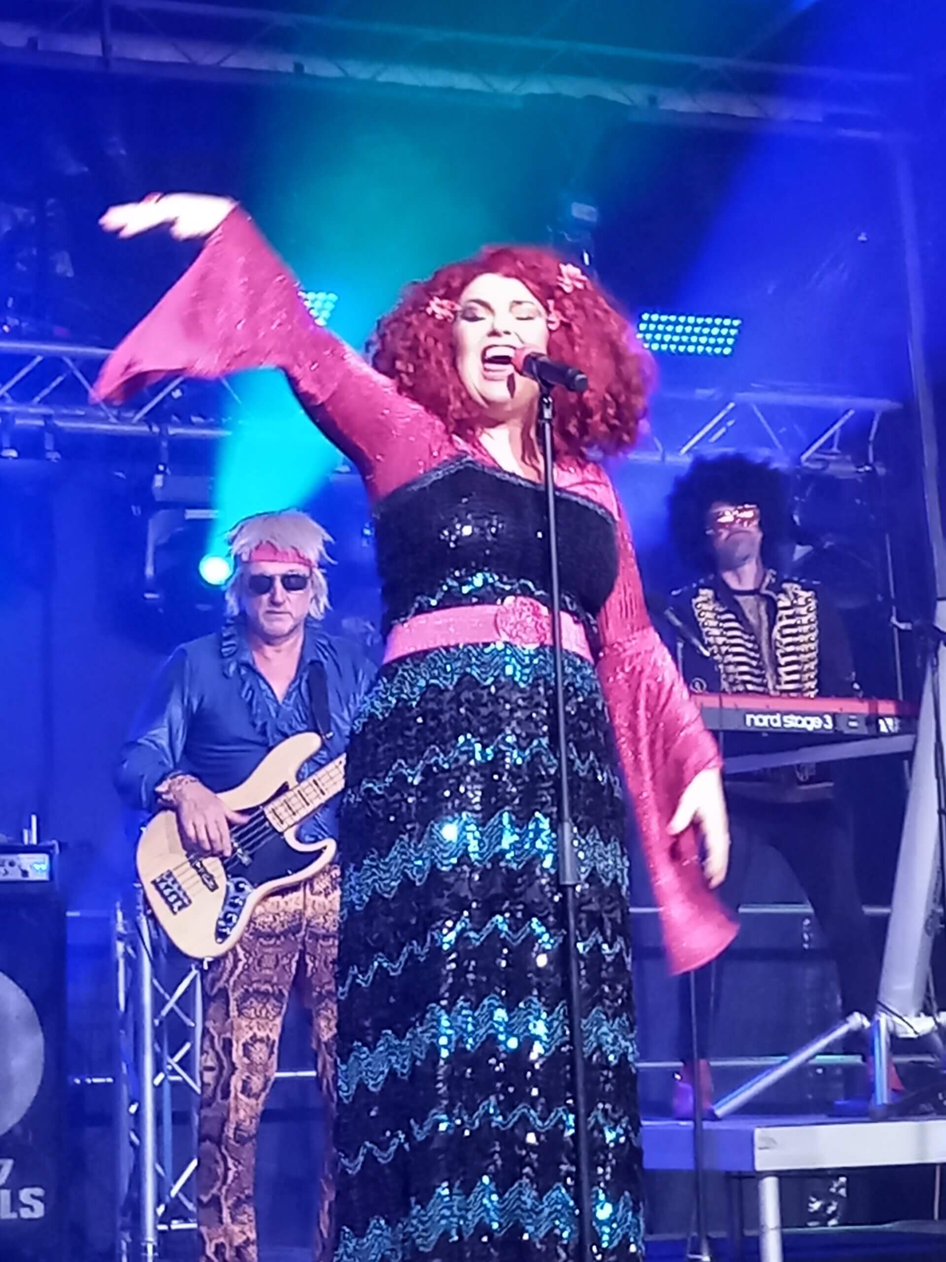 Frau mit rotem Lockenhaar singt leidenschaftlich auf der Bühne, begleitet von Band in retro-inspirierten Outfits.