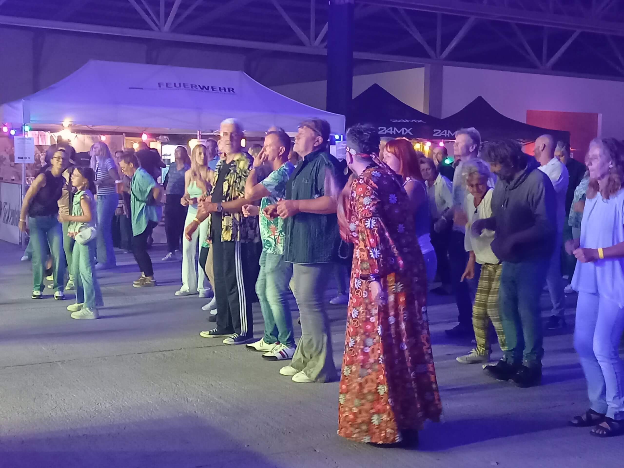 Gruppe von Menschen tanzt fröhlich bei einem Event im Freien, nachts unter bunter Beleuchtung, Zelte im Hintergrund.