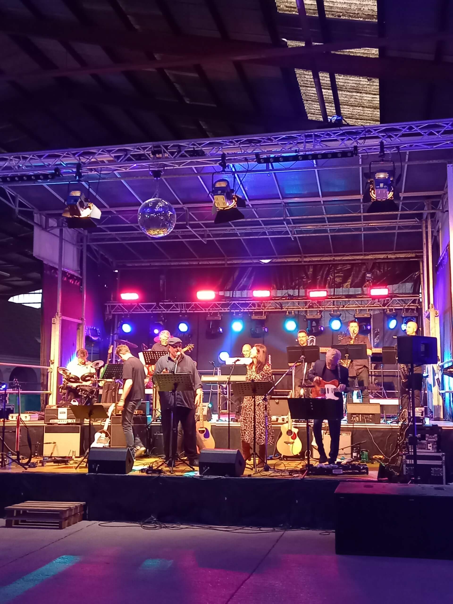 Live-Band-Performance auf einer bunt beleuchteten Bühne in einer Halle mit verschiedenen Instrumenten und Sängern.