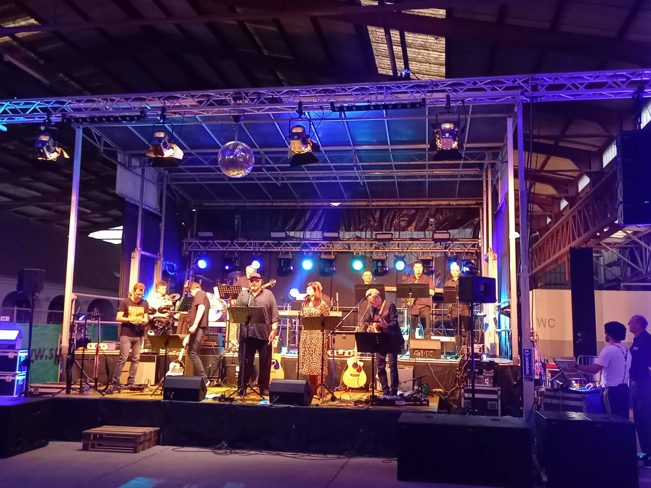 Live-Konzert in einer Halle, Musiker auf beleuchteter Bühne mit Instrumenten und Mikrofonen.