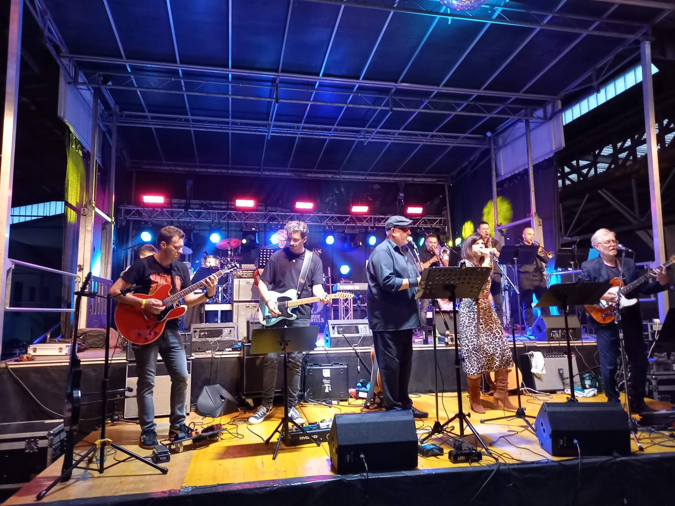 Band spielt Live-Konzert auf Bühne mit Gitarren und Bläsern unter bunten Scheinwerfern. Stimmungsvoll und energiegeladen.