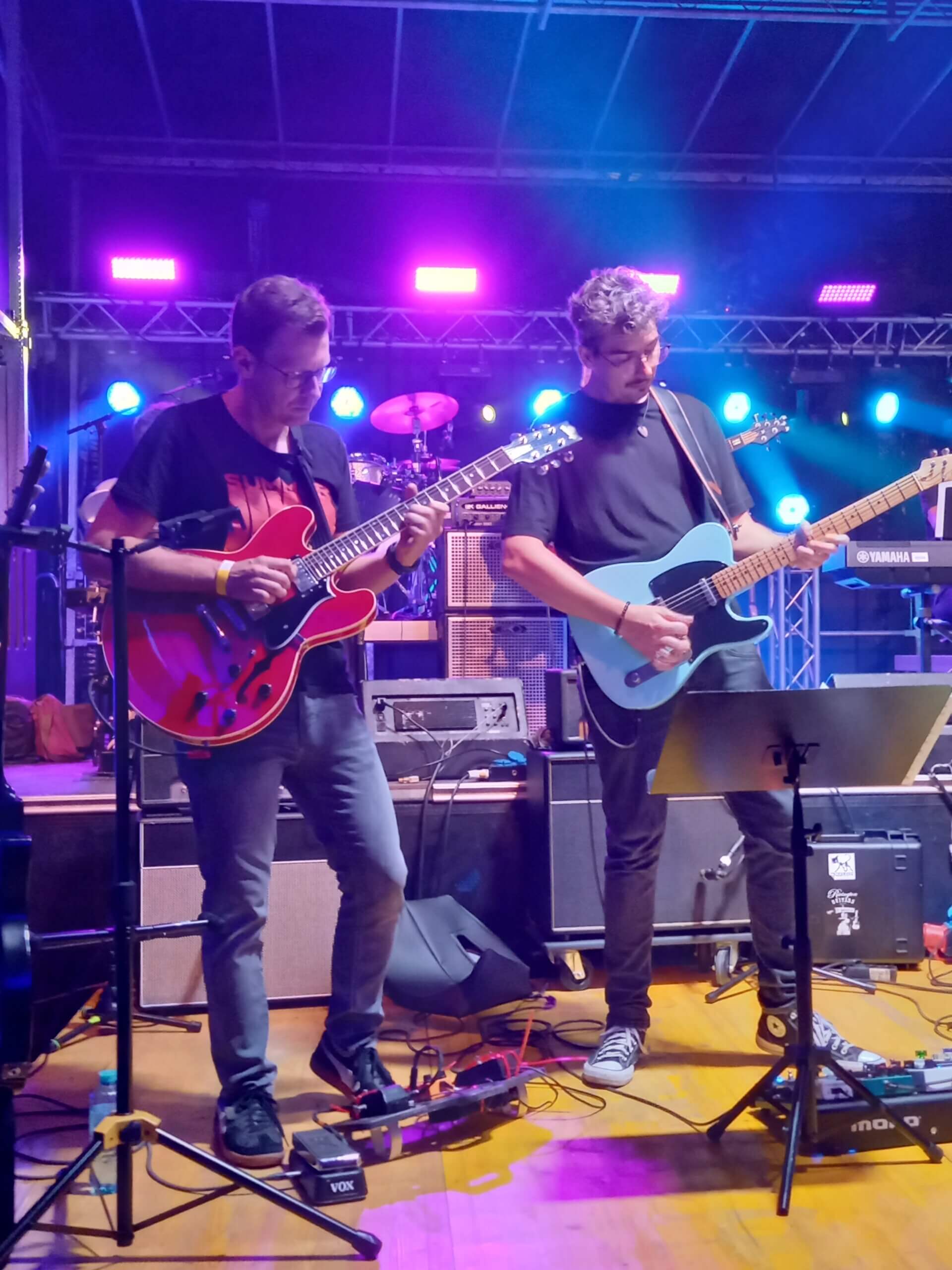 Zwei Gitarristen auf einer Bühne während eines Live-Auftritts mit bunten Lichtern im Hintergrund.