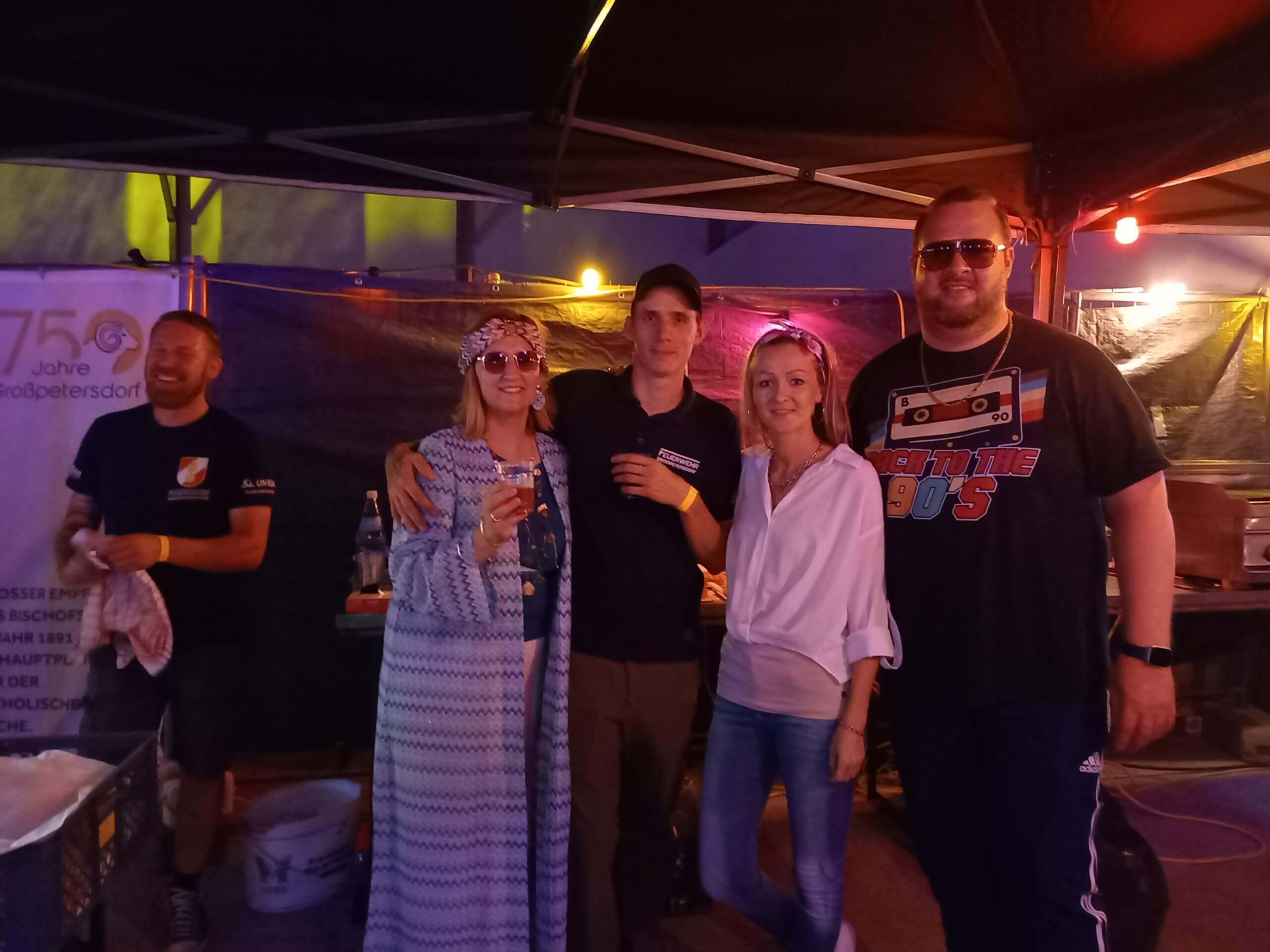 Gruppe feiert ein Festival mit Retro-Kleidung unter einem Partyzelt bei Nacht. Stimmung: fröhlich und entspannt.