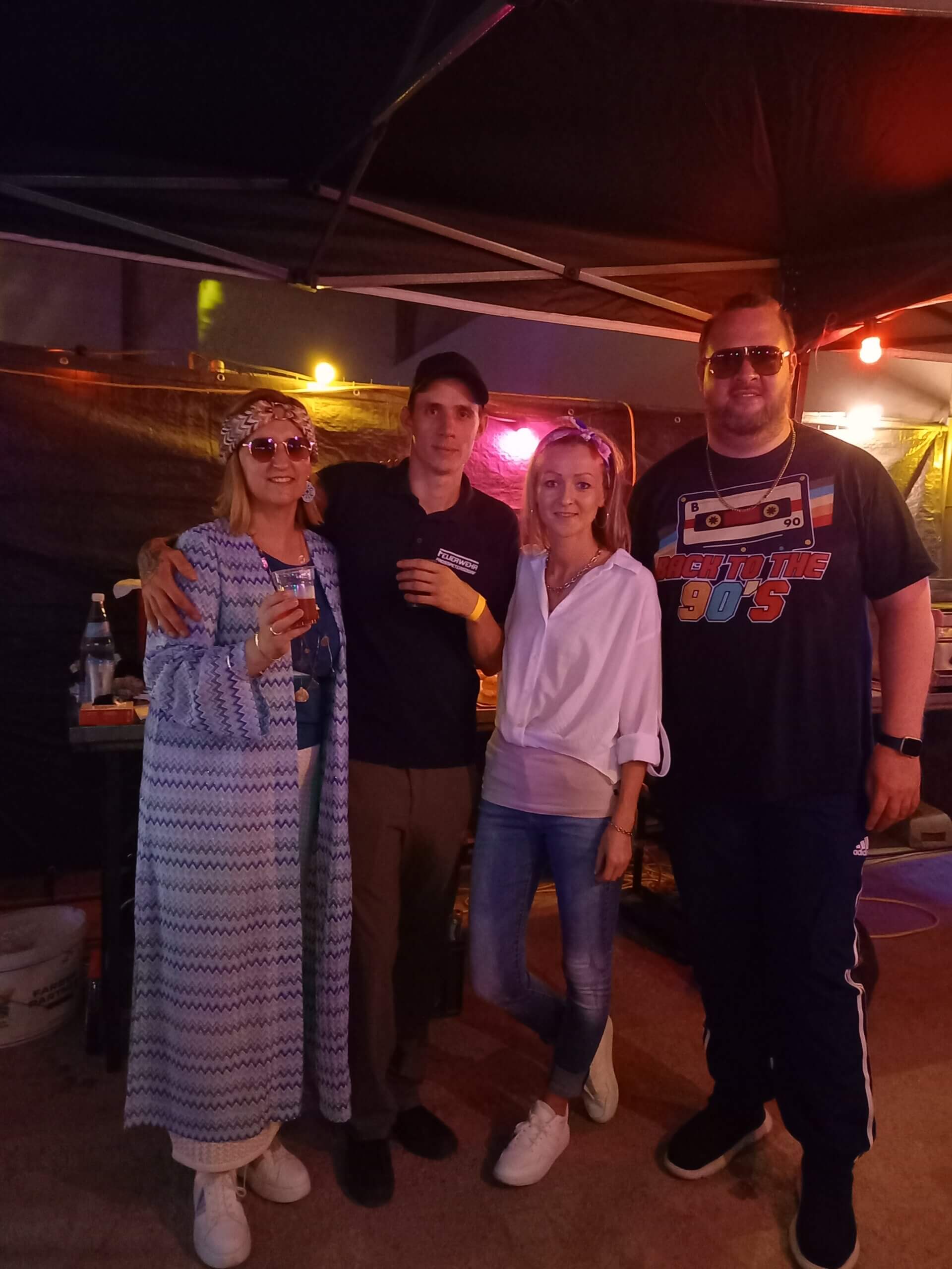 Vier Menschen posieren fröhlich auf einer 90er-Jahre-Party mit bunten Lichtern im Hintergrund.