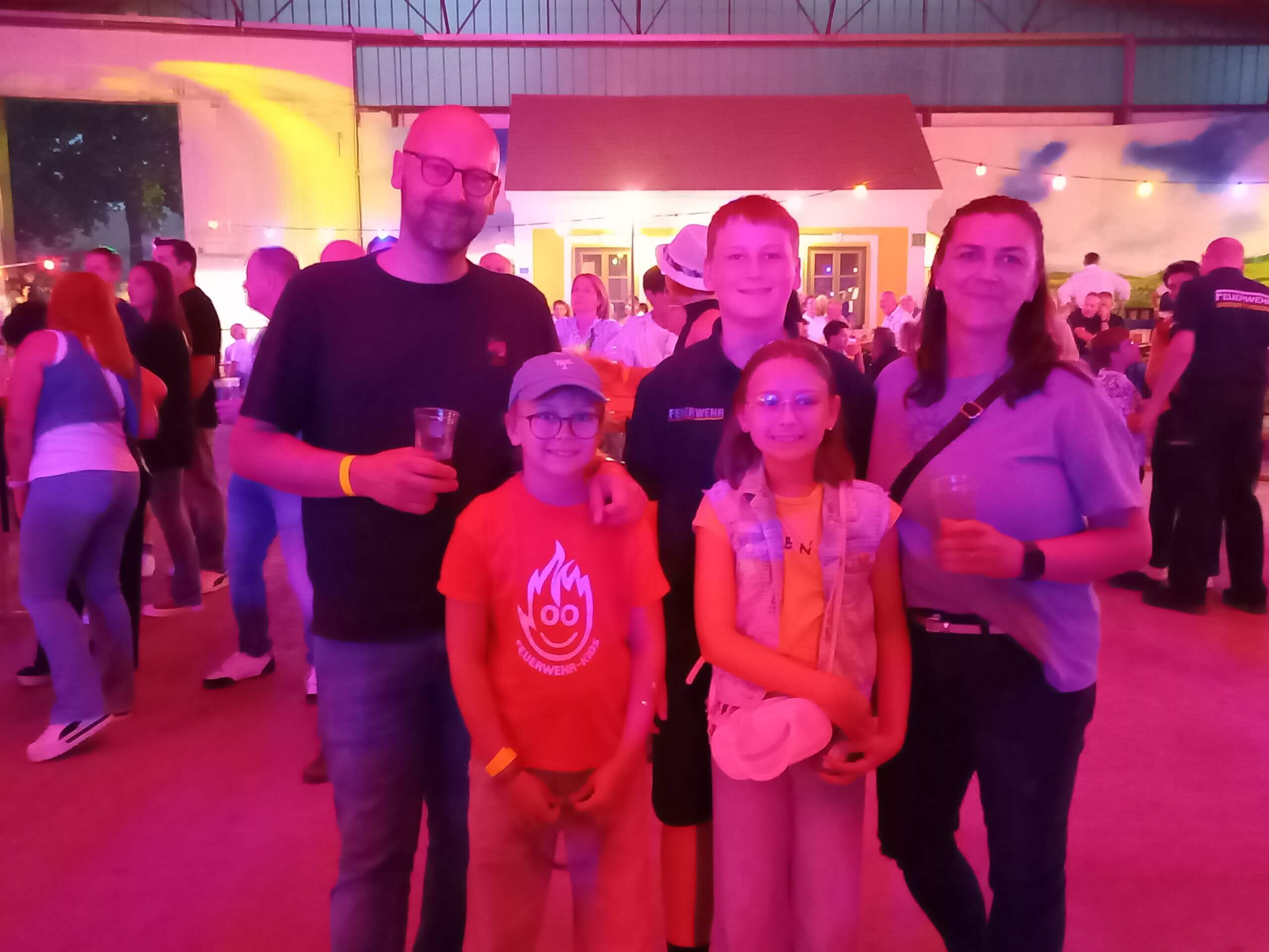 Familie bei einem fröhlichen Event in einer bunt beleuchteten Halle mit anderen Besuchern im Hintergrund.