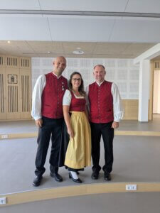 Drei Personen in traditioneller Tracht, lächelnd vor einer modernen Innenraumkulisse.
