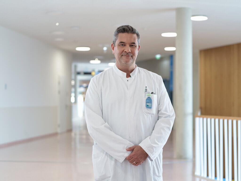Arzt, Leiter der Kardiologie, im weißen Kittel steht lächelnd im Krankenhausflur, professionell und vertrauensvoll.