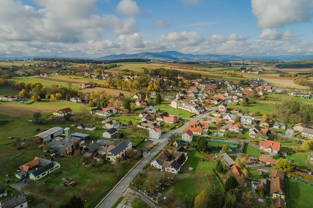 Rohr bei Hartberg_©Karl Schrotter Luftaufnahme eines idyllischen Dorfes inmitten grüner Landschaft und sanfter Hügel an einem sonnigen Tag.