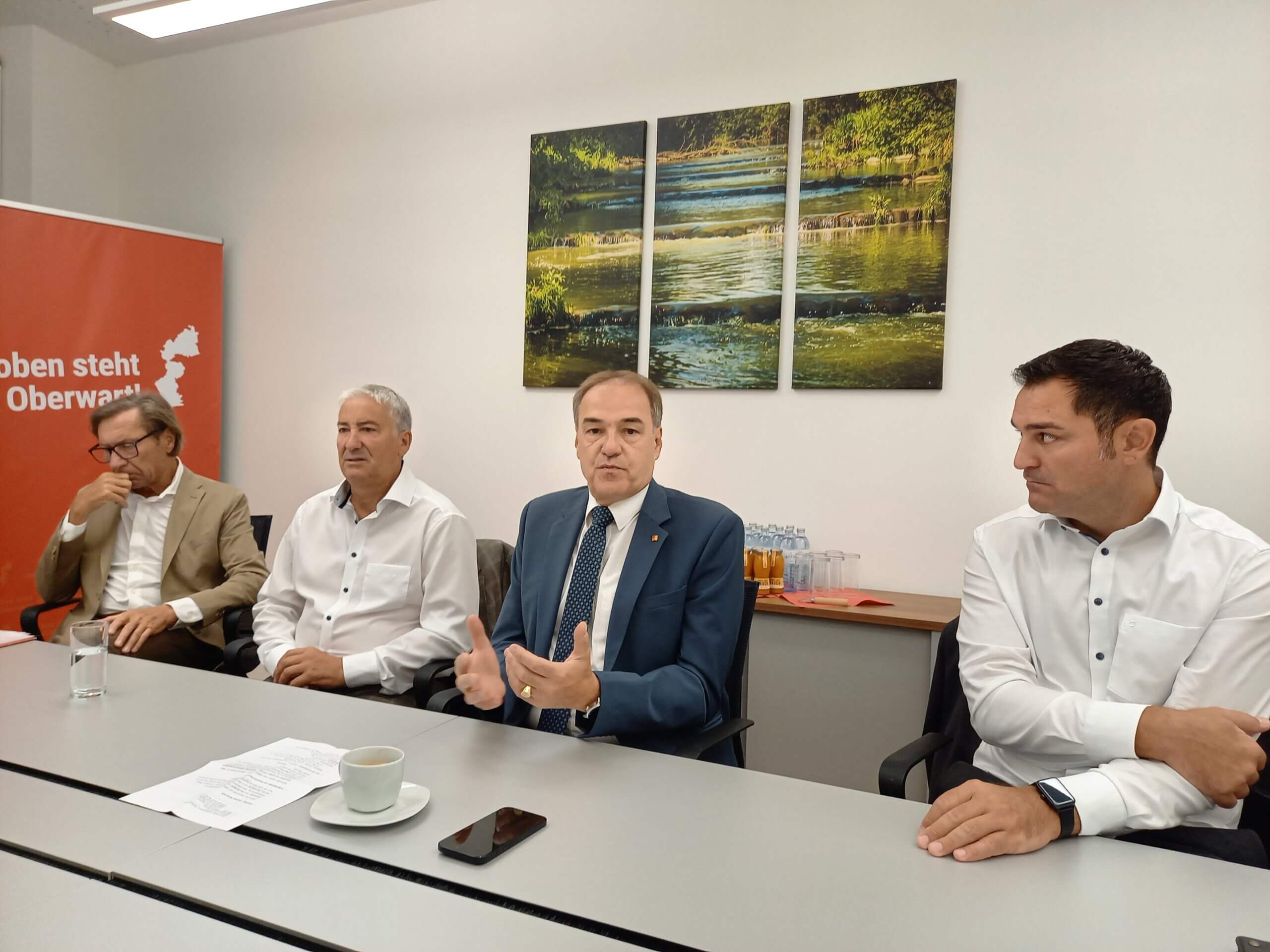 Vier Männer in Businesskleidung diskutieren in einem Konferenzraum; im Hintergrund ist ein Naturbild zu sehen.