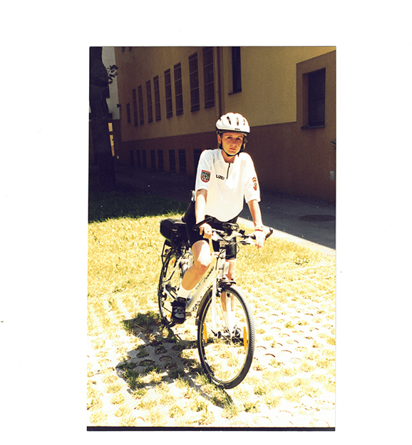 Sandra Polizei Fahrraduniform 1999 Person auf einem Fahrrad mit Helm auf sonniger Wiese vor einem Gebäude, ideal für urbane Fahrradtouren.