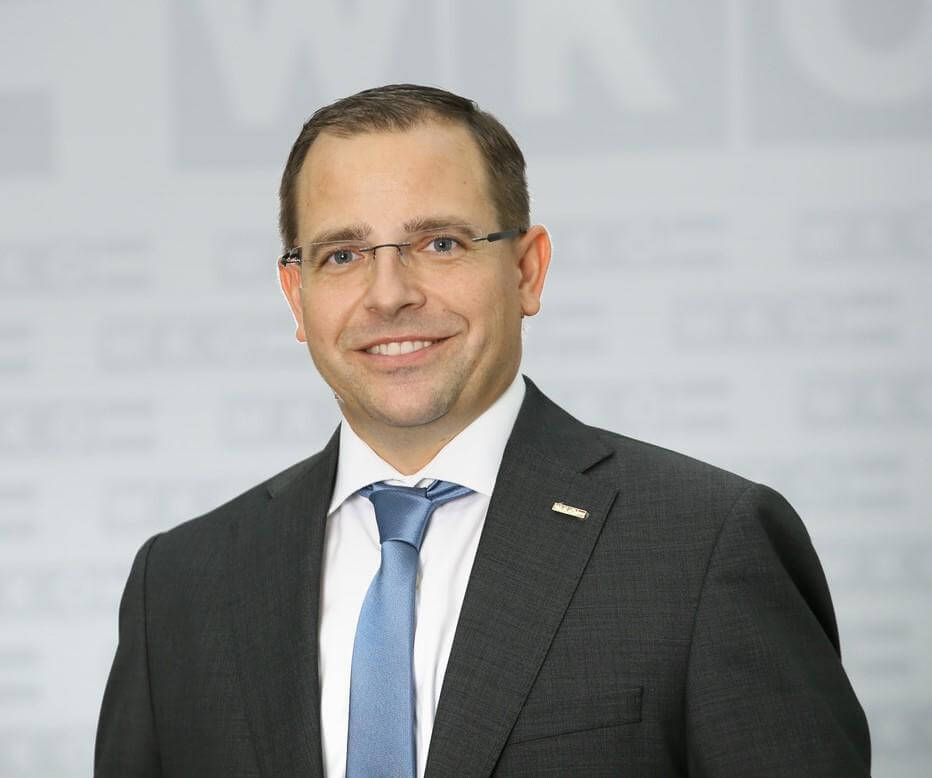 Ein Mann in Anzug und Krawatte lächelt professionell in die Kamera vor einem neutralen grauen Hintergrund.