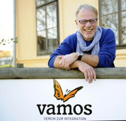image_79ce2402f5aed4eeb311853a7c98b4e0_255x243 Lächelnde Person lehnt an einem Schild mit vamos Logo, Verein zur Integration, im Hintergrund ein Gebäude.