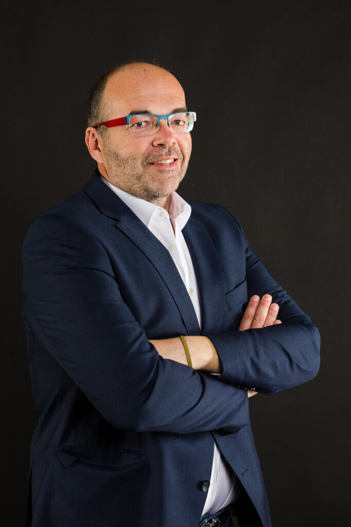 zVg Mann im Anzug mit Brille vor schwarzem Hintergrund, verschränkte Arme, selbstbewusste Haltung.
