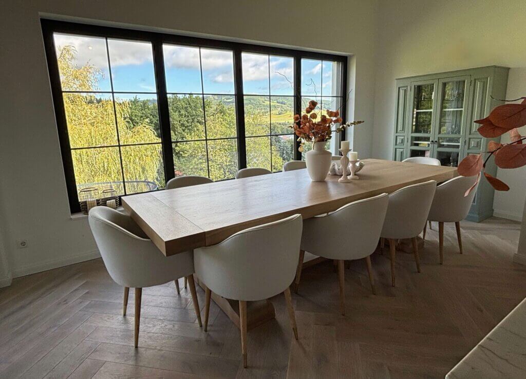 Moderner Essbereich mit großem Holztisch und weißen Stühlen, Blick auf die Natur durch große Fenster.