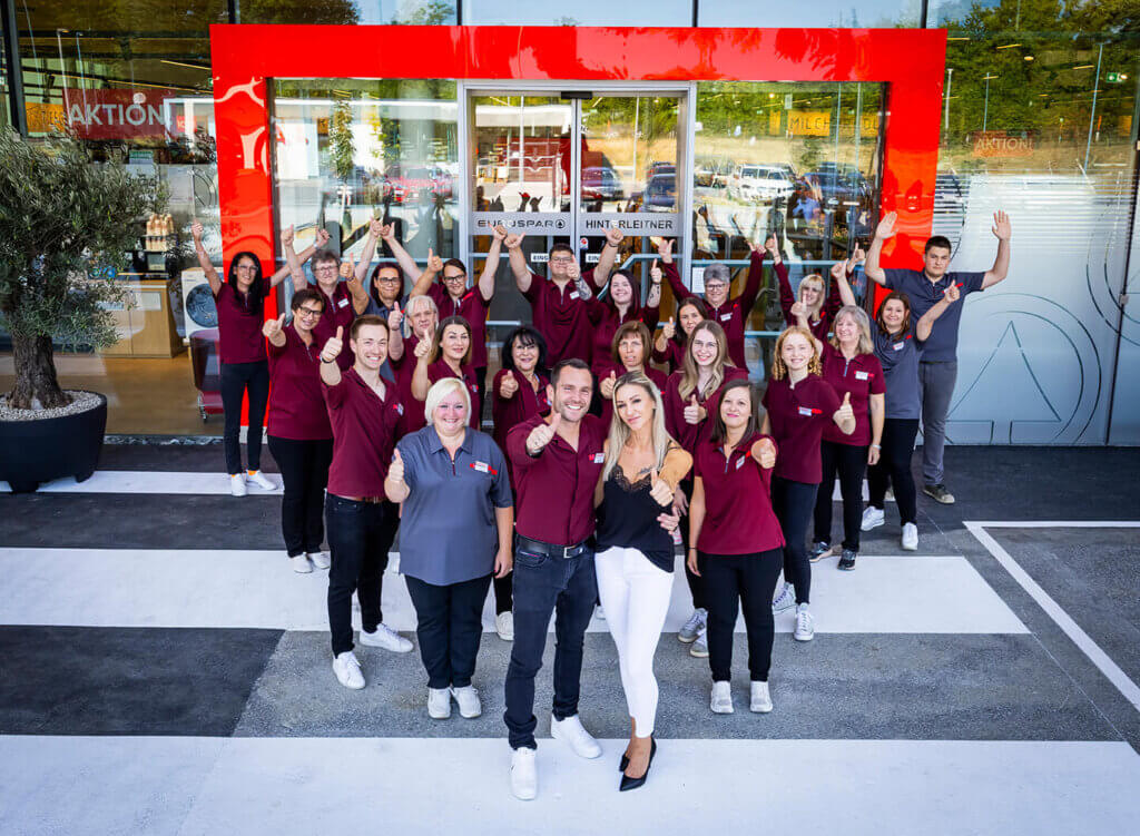Mitarbeiter eines Supermarkts jubeln vor dem Eingang in roten Uniformen, Daumen hoch, Teamspirit, Kundenzufriedenheit.