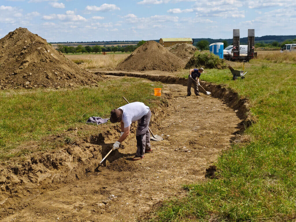 Archäologen graben auf einer grünen Wiese bei einer Ausgrabungsstätte; Erdarbeiten im Gange.