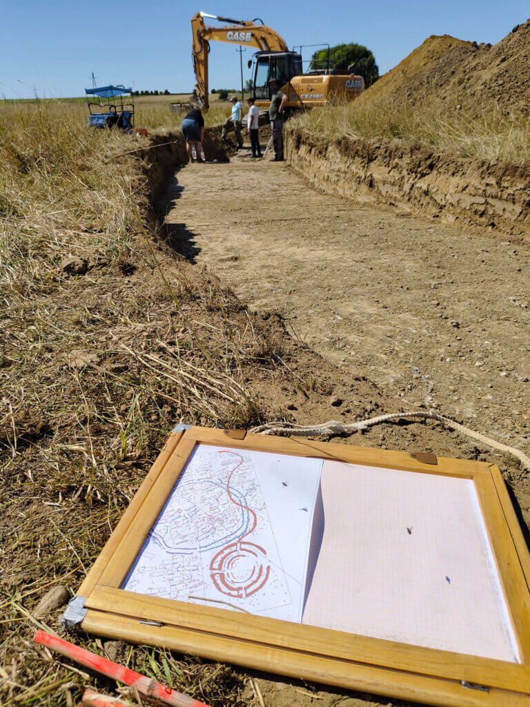 Archäologische Ausgrabung mit Bagger und Lageplan im Vordergrund auf einer Baustelle.