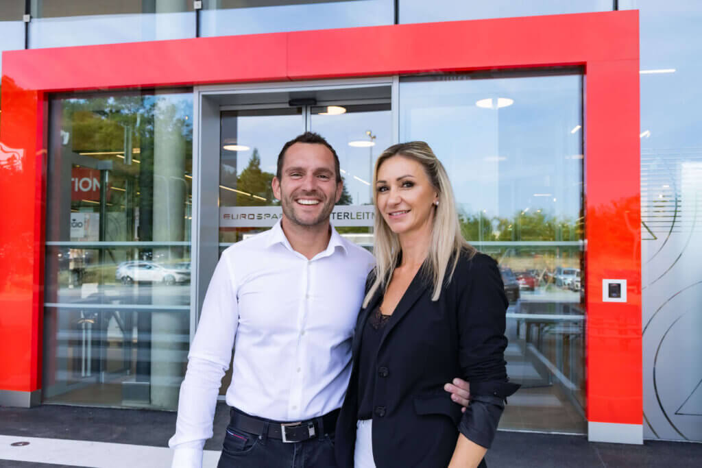 Zwei lächelnde Personen vor einem modernen Bürogebäude mit rotem Rahmen, Firmenlogo im Hintergrund.
