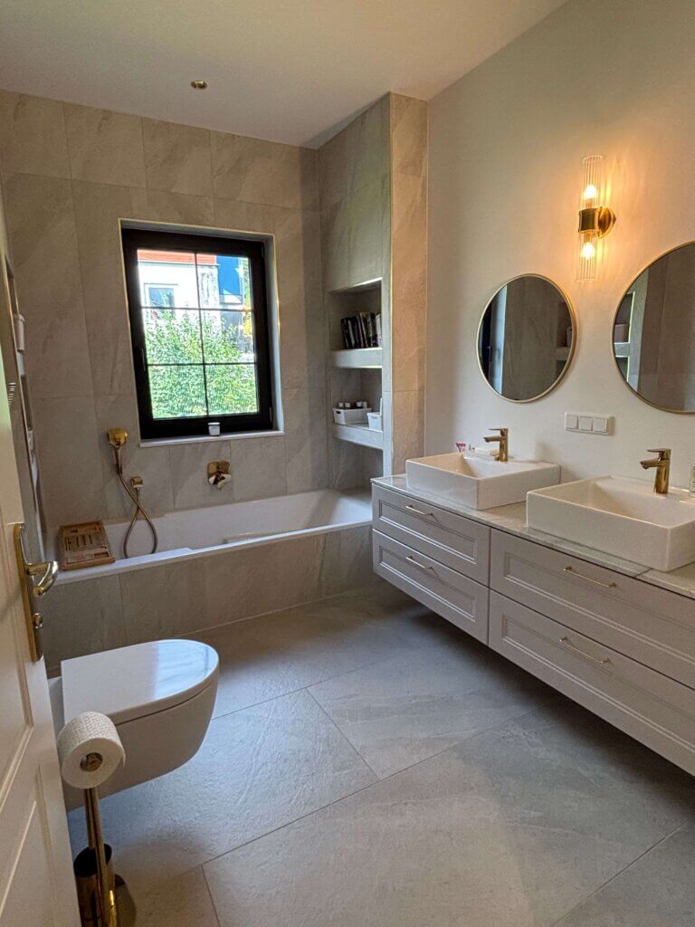 Modernes Badezimmer mit Badewanne, Doppelwaschbecken und großen Fliesen, beleuchtet durch stilvolle Wandlampen.