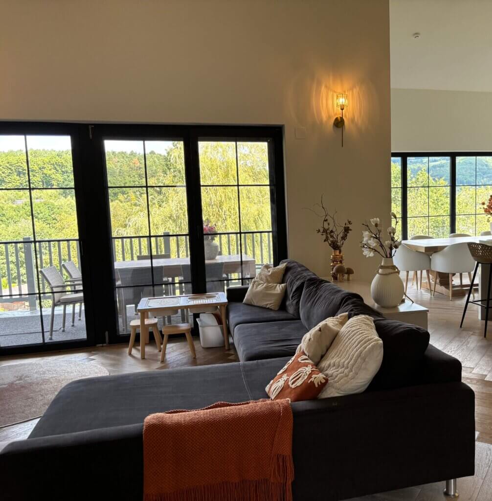 Modernes Wohnzimmer mit großem Sofa, Aussicht auf Terrasse und grüne Landschaft durch große Fenster. Rustikal und hell.
