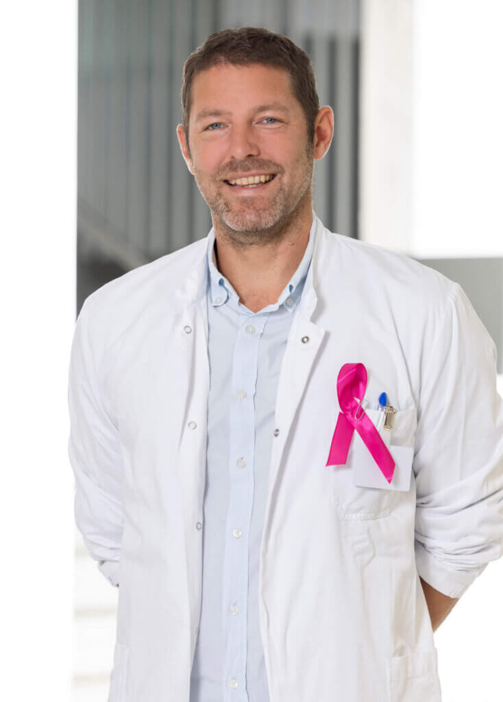 Ein lächelnder Arzt mit rosa Schleife zeigt Unterstützung für Brustkrebsbewusstsein in einer modern aussehenden Umgebung.
