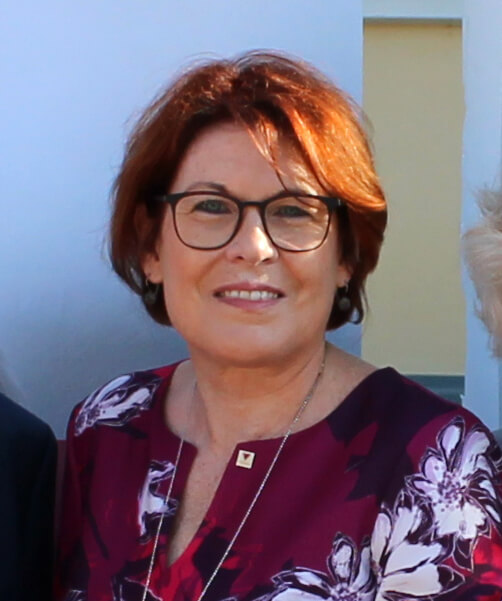 Eine Person mit braunem Haar und Brille lächelt in die Kamera, trägt ein gemustertes Oberteil.