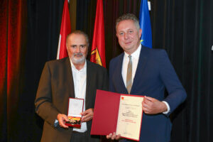 Zwei Männer bei Preisverleihung mit Urkunde und Medaille, vor farbigen Fahnen auf der Bühne.